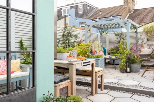 un giardino pavimentato con fioriere rialzate dipinte di azzurro e pergola, pavimentazione esagonale, sedia e poggiapiedi in rete grigia e recinzione a doghe grigio chiaro